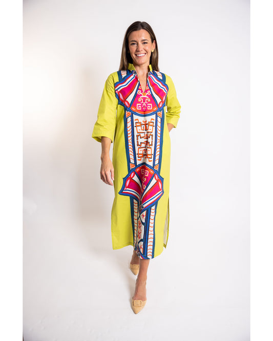 A woman modeling a bright yellow kaftan dress with a large, colorful geometric and tribal-style embroidered pattern in pink, blue, and white down the front and on the chest. She is standing against a plain white studio background.