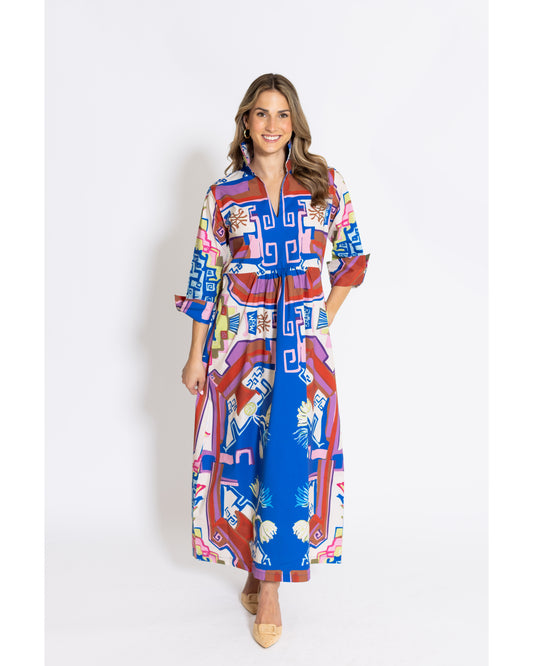 A woman modeling a long-sleeved maxi shirt dress with a vibrant geometric and tropical print. The dress features a bold mix of royal blue, red, pink, yellow, and white patterns, including palm leaves and geometric shapes.