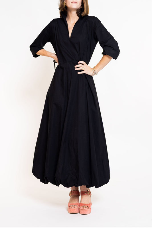 A woman modeling a black maxi dress with three-quarter sleeves, a wrap-style V-neckline, and a self-tie waist belt, paired with pink platform sandals against a plain white studio background.
